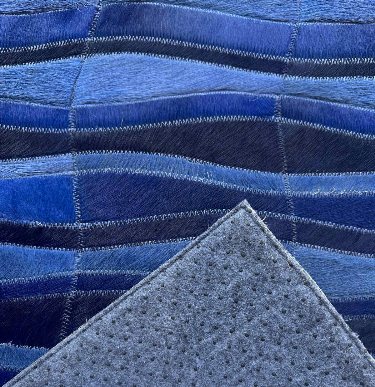 Cavo stitched cowhide rug sample with a corner turned back to reveal gray felt backing and small black rubber grip dots.