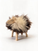 A small sheep-shaped figurine with a fluffy fur body in cream and dark brown, standing on four wooden legs against a plain white background.