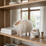 A fluffy white sheep figurine with light wood legs and ears stands on a wooden shelf beside stacked books, a small gray vase, a white orb, and a brass candlestick, with a softly blurred window and curtains in the background.