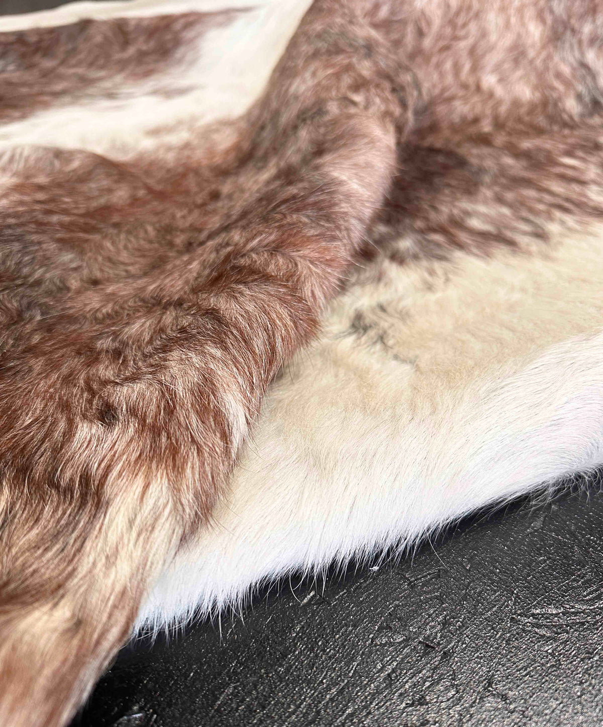Close-up of a brown and white cowhide fur rug, showing the texture of the hair and the natural color variation, laid over a dark textured surface.