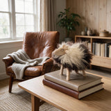 A fluffy sheep figurine with cream and dark brown fur stands on a stack of two books on a wooden table in a cozy living room setting. A brown leather armchair with a gray knit blanket is visible in the background near a window.